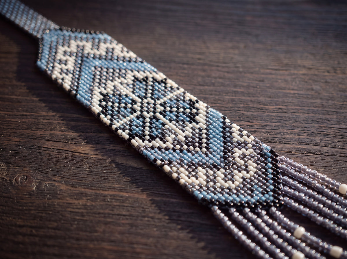 Close-up detail of a handmade beaded necklace strap showing blue, black and white woven geometric pattern with beaded fringe on dark wood