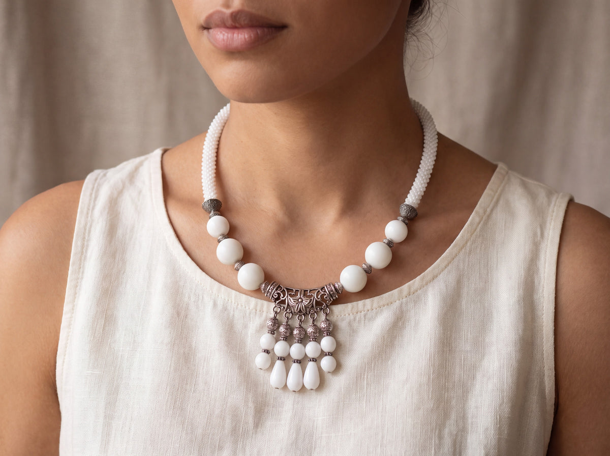 Handmade white beaded necklace with large glossy beads, ornate silver-tone filigree pendant and teardrop dangles, worn on a model at the collarbone