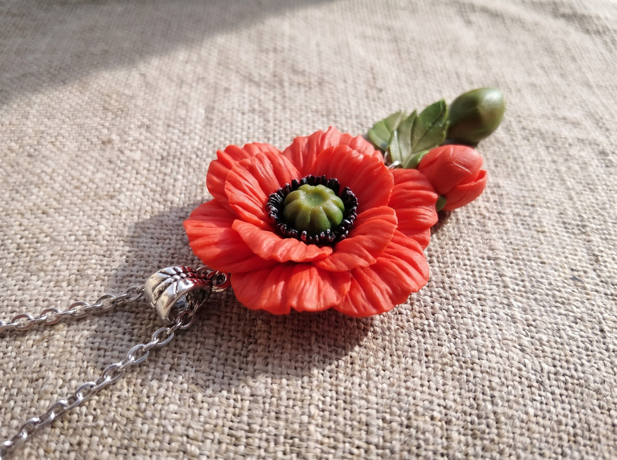Close-up of a handmade red poppy flower pendant with sculpted petals, bud, and green leaves on a silver chain, laid on woven linen.