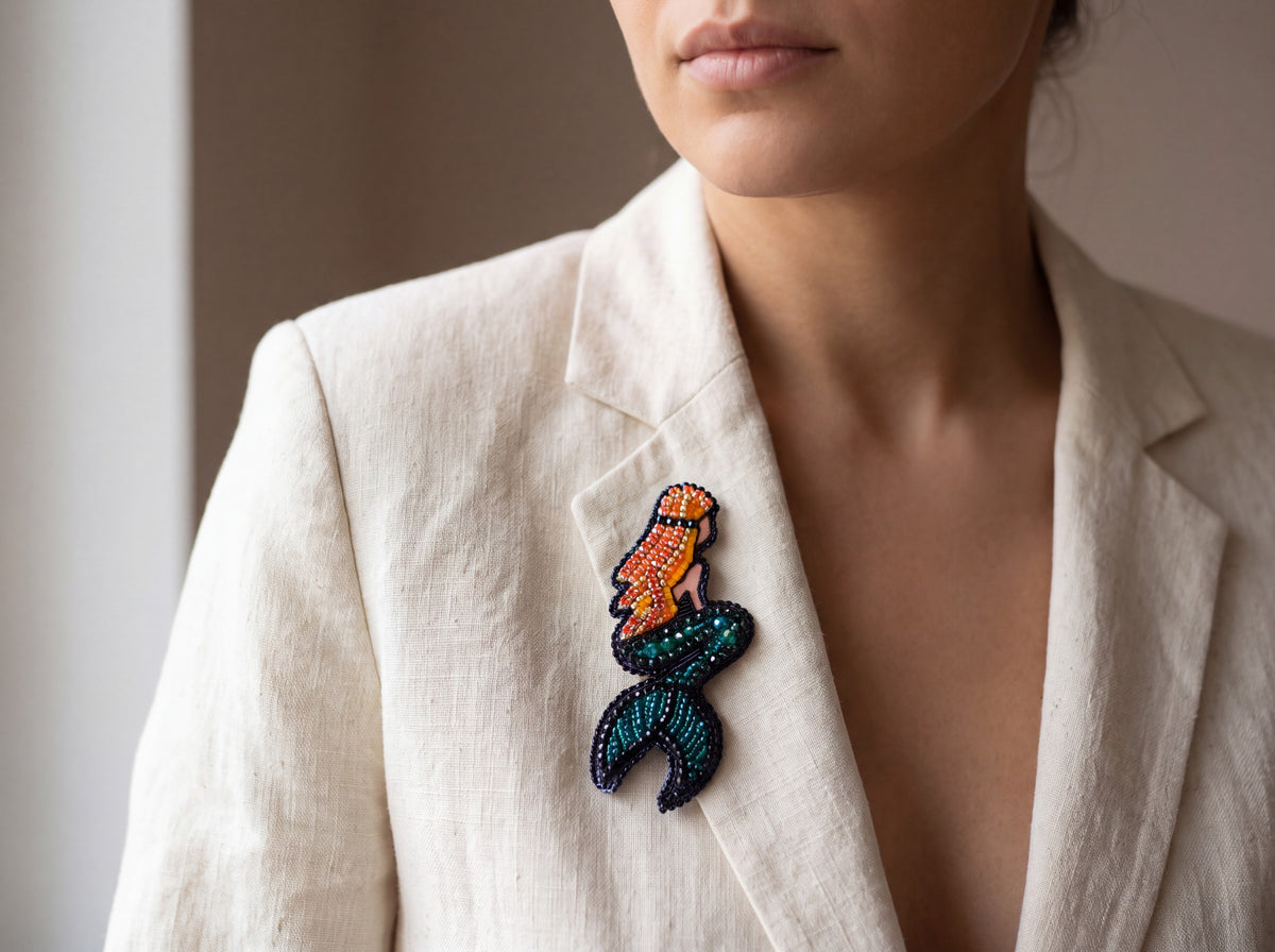 Handmade bead-embroidered mermaid brooch with orange beaded hair and teal tail, pinned to the lapel of a cream linen blazer worn by a model.