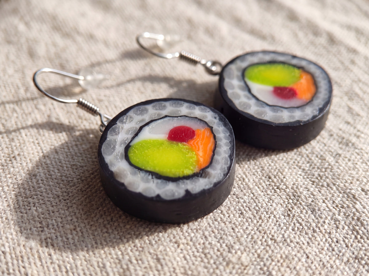 Close-up of handmade polymer clay maki sushi roll earrings with realistic rice, nori, avocado and salmon detail lying on linen fabric