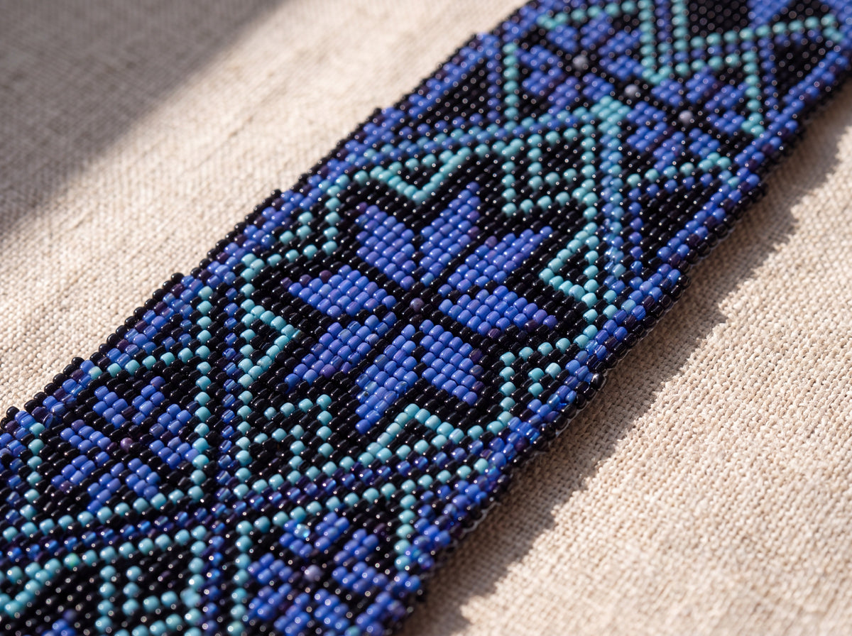 Close-up detail of a handmade beaded tie showing woven blue, turquoise and black seed beads forming geometric eight-point stars on linen