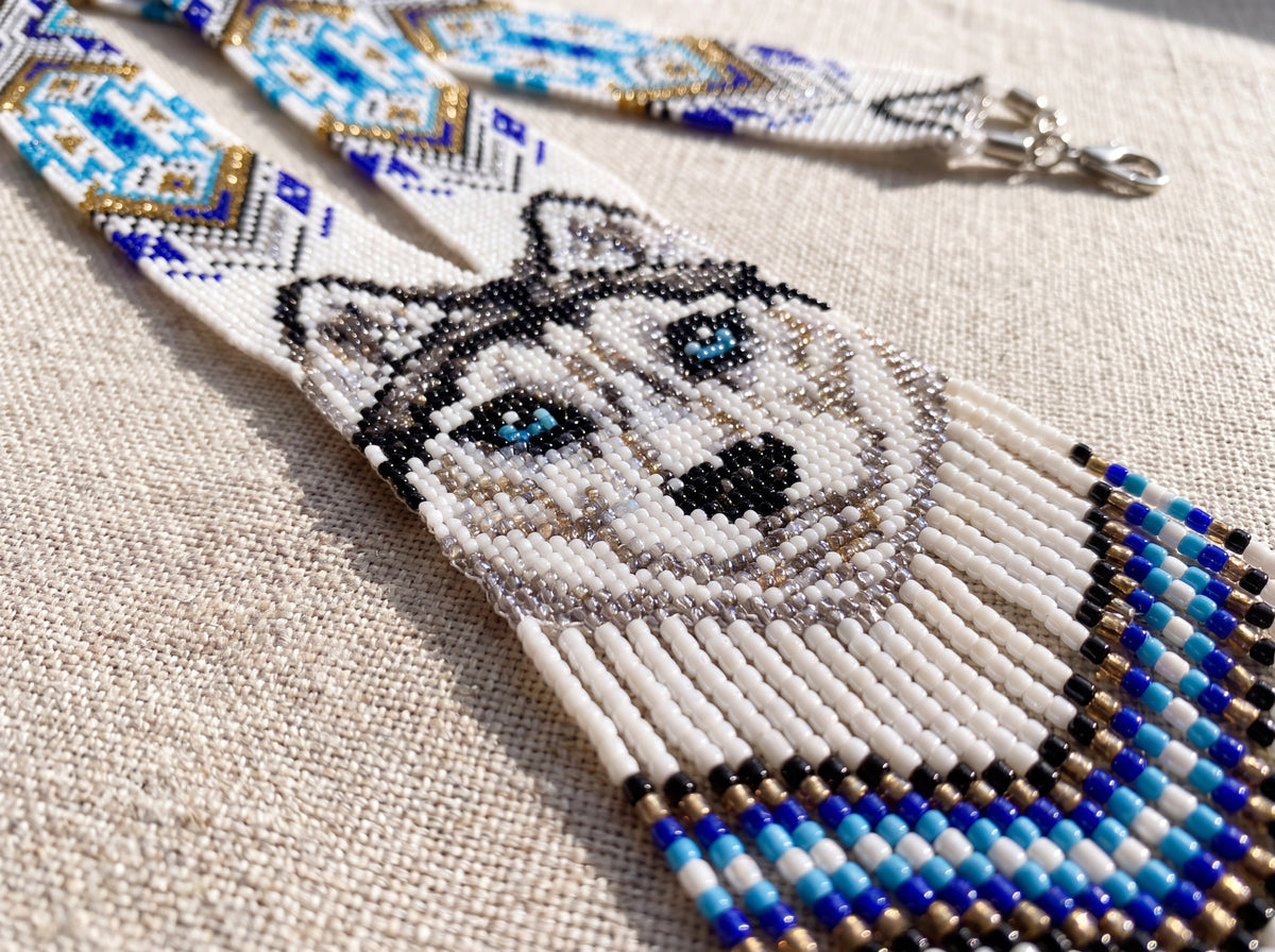 Close-up of a handmade beaded wolf necklace showing the blue-eyed wolf face in seed beads with blue, white and gold fringe on linen fabric