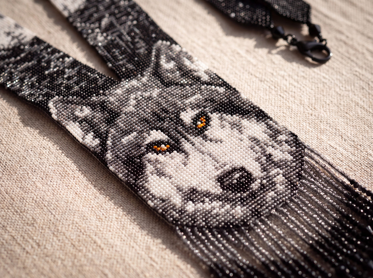 Close-up detail of a handmade beaded wolf gerdan showing the amber-eyed gray wolf face woven in seed beads with dark beaded fringe