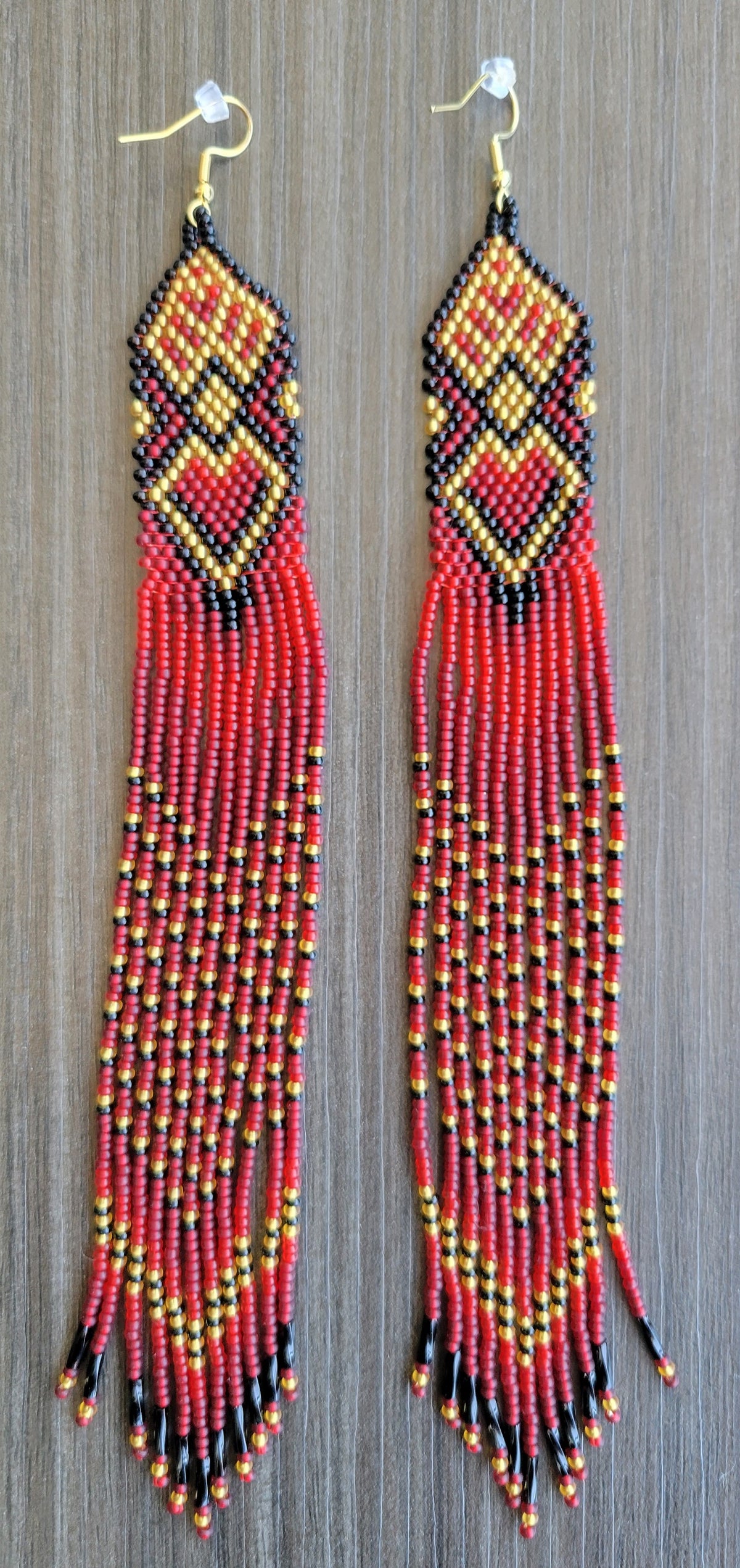 Handmade extra long beaded fringe earrings in red, gold and black with heart motif on dark wood