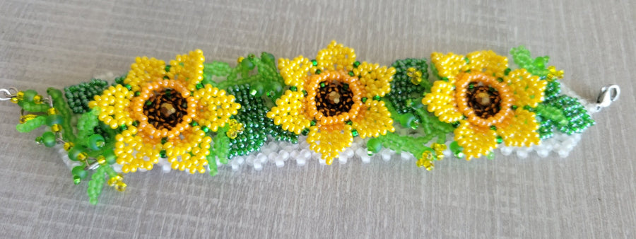 Beaded flower bracelet with three yellow sunflowers and green leaves on a white seed bead band, flat lay