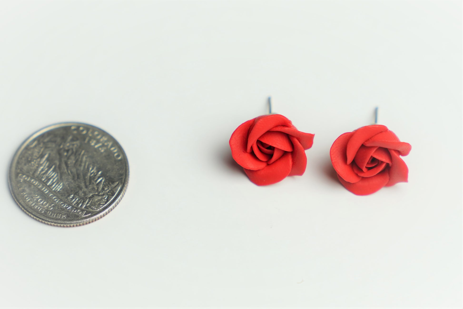 Red rose stud earrings next to a quarter coin for size comparison, flat lay on white