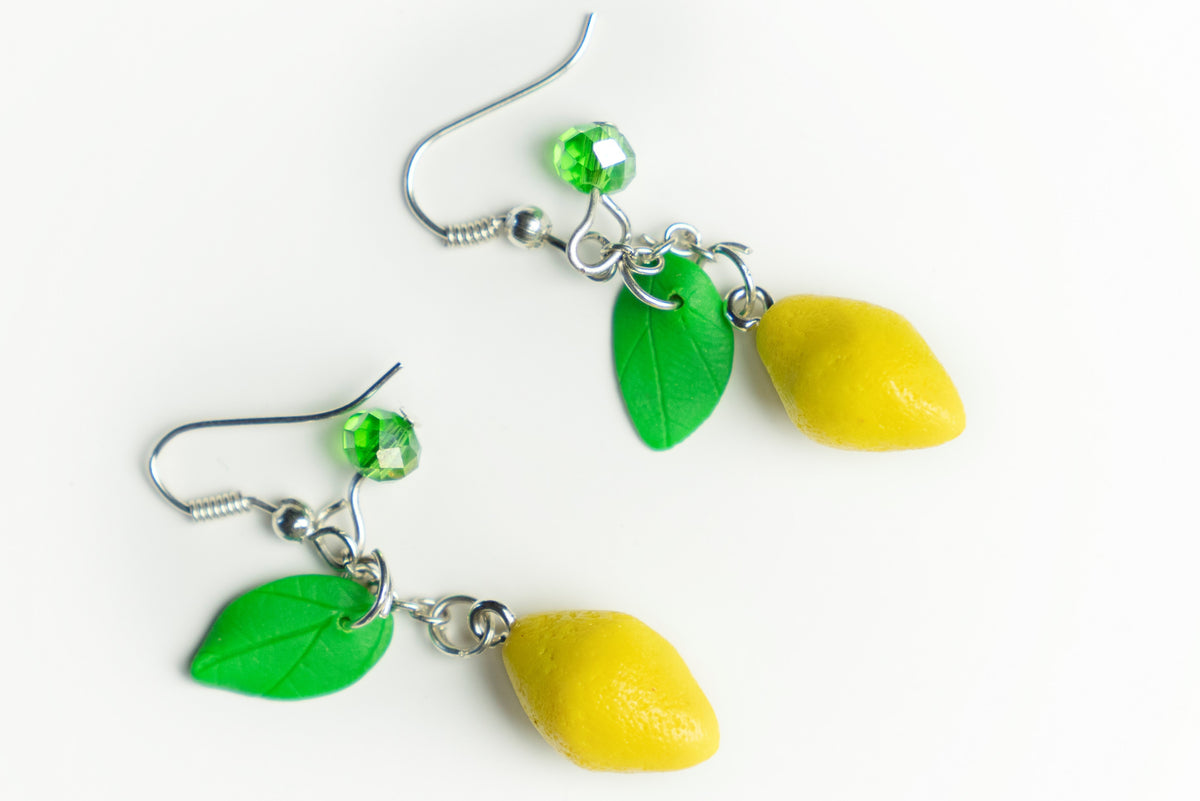 Pair of yellow lemon dangle earrings in polymer clay with leaves and green crystal beads, angled flat lay