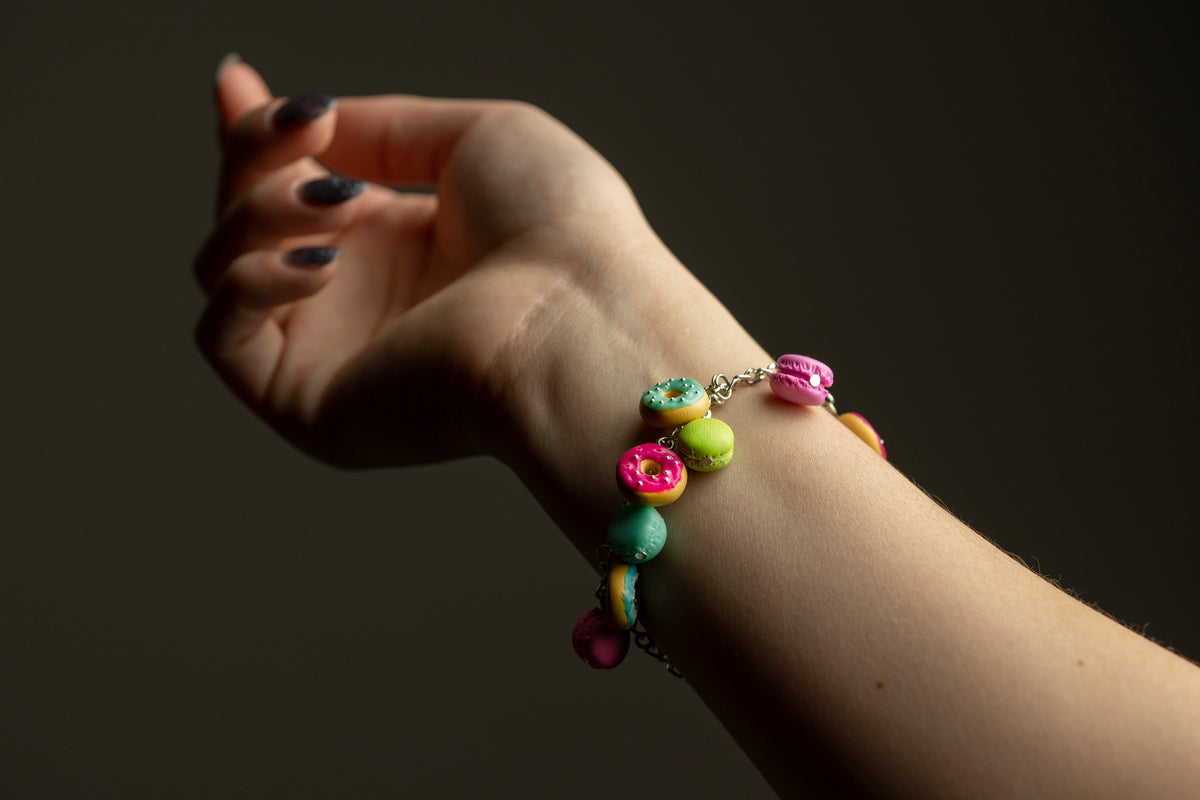 Polymer clay donut and macaron charm bracelet worn on a raised wrist against a dark background