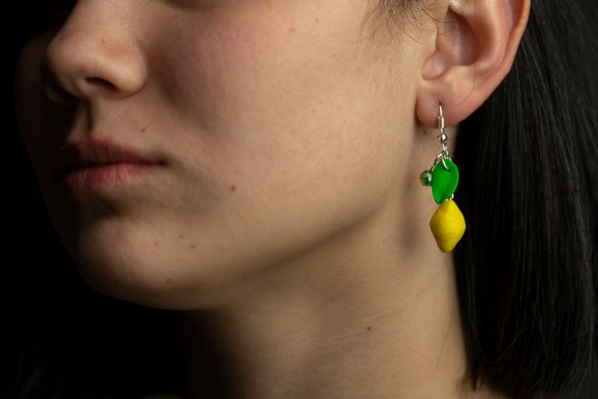 Polymer clay lemon earring with leaf and green crystal worn on a model's ear against a dark background