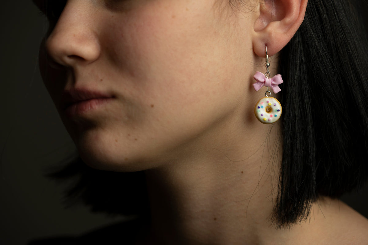 Polymer clay donut earring with pink bow worn by a model, close-up at the ear against dark hair
