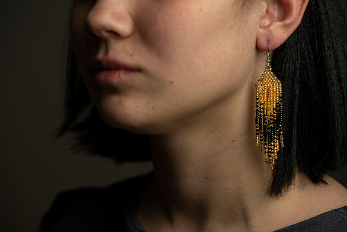 Handmade black and gold beaded fringe earring worn by a model with dark hair, dramatic lighting